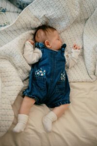 A sleeping newborn girl in a denim dress on a bed with blue and white sheets in Littleton, Massachusetts.