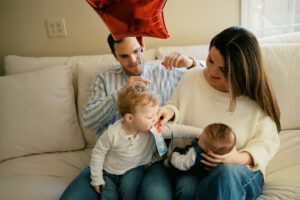 A family plays at home in their living room near Acton, Massachusetts. They sit on a white couch, the mother holding a newborn girl while a toddler eats a snack and reaches for his sister's face. The father holds a red balloon.