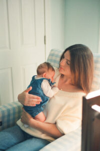 A mother in a white sweater and jeans holds her newborn daughter in a denim dress. They are a sitting in a blue gingham recliner in a nursery near Acton, Massachusetts.