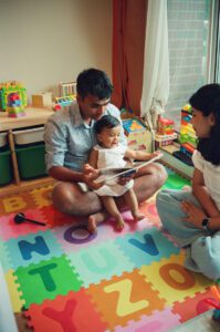 A father plays with his one year old daughter on a colorful play mat on the floor of their apartment in Cambridge, Massachusetts, while the mother looks on. Captured on 35mm film.
