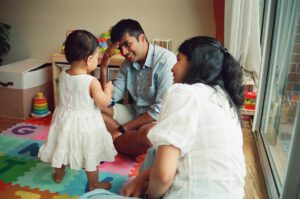 A father smiles at his one year old daughter as the mother looks on from the floor of their apartment in Cambridge, Massachusetts. Captured on 35mm film.