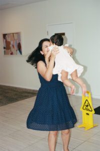 A mother in a dark blue dress holds her one-year-old daughter in a white dress and twirls her around the lobby of their apartment building in Cambridge, Massachusetts. Captured on 35mm film.