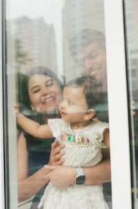 A view through a window of a mother in a dark blue dress holding her one-year-old daughter in a white dress while the father smiles down at them. They are in the lobby of their apartment building in Cambridge, Massachusetts. Captured on 35mm film.