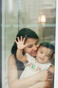 A view through a window of a mother in a dark blue dress cuddling her one-year-old daughter in a white dress in the lobby of their apartment building in Cambridge, Massachusetts. Captured on 35mm film.