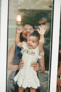 A view through a window of a mother in a dark blue dress holding her one-year-old daughter in a white dress while the father stands behind them. They are in the lobby of their apartment building in Cambridge, Massachusetts. Captured on 35mm film.