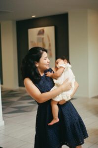 A mother in a dark blue dress holds her one-year-old daughter in a white dress in the lobby of their apartment building in Cambridge, Massachusetts. Captured on 35mm film.