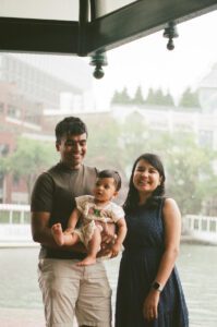 A mother, father, and a toddler girl in a white dress stand under a pavilion by the Charles river in Cambridge, Massachusetts while rain pours down behind them. Captured on 35mm film.