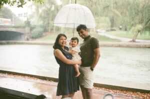 A family with a toddler girl in a white dress stands under a clear umbrella on the Charles river in Cambridge, Massachusetts while rain pours down behind them. Captured on 35mm film.