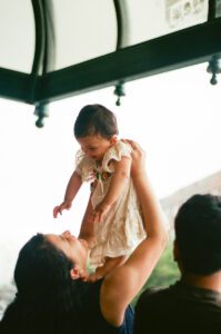 A mother, lifts her one-year-old daughter in a white dress as they sit on a bench by the Charles river in Cambridge, Massachusetts. Captured on 35mm film.
