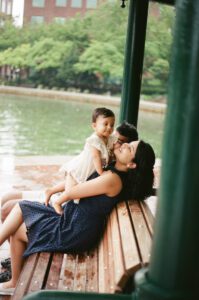 A mother in a dark blue dress holds her one-year-old daughter in a white dress as they sit on a bench in Cambridge, Massachusetts. The father sits behind them while rain pours down. Captured on 35mm film.