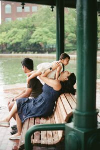 A mother in a dark blue dress lifts her one-year-old daughter in a white dress as they sit on a bench in Cambridge, Massachusetts. The father sits behind them while rain pours down. Captured on 35mm film.