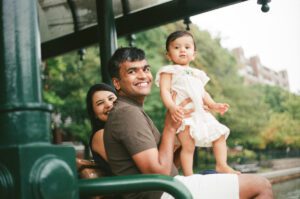 A father in a brown shirt holds his one-year-old daughter in a white dress on his lap as they sit on a bench in Cambridge, Massachusetts. The mother sits behind them and smiles while rain pours down. Captured on 35mm film.