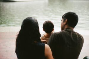 View of a family from behind as they sit near the Charles river in Cambridge, Massachusetts. The toddler girl sits between her mother and father.