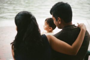 View of a family from the side as they sit near the Charles river in Cambridge, Massachusetts. The toddler girl sits between her mother and father.