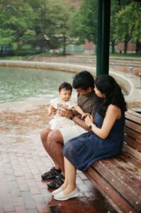 A father in a brown shirt holds his one-year-old daughter in a white dress on his lap as they sit on a bench in Cambridge, Massachusetts. The mother in a dark blue dress sits next to them while rain pours down around them. Captured on 35mm film.