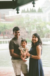 A mother, father, and a toddler girl in a white dress stand under a pavilion laughing by the Charles river in Cambridge, Massachusetts while rain pours down behind them. Captured on 35mm film.