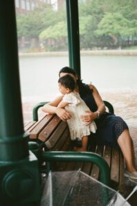 A mother in a dark blue dress holds her one-year-old daughter in a white dress they sit on a bench by the Charles River in Cambridge, Massachusetts and watch the rain pour down around them. Captured on 35mm film.
