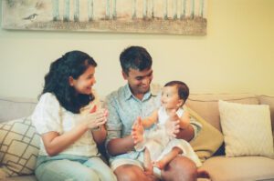 A mother and father hold their one year old daughter between them and smile at her as she claps on the couch in their apartment in Cambridge, Massachusetts. Captured on 35mm film.