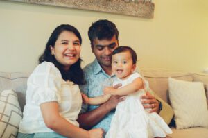 A mother and father hold their one year old daughter between them and smile at the camera on the couch in their apartment in Cambridge, Massachusetts. Captured on 35mm film.