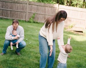 Family of four plays in their backyard in Greater Boston. The mother reaches down to a toddler boy, both wearing white, and the father in a blue striped shirt holds a newborn girl. Captured on 35mm film.