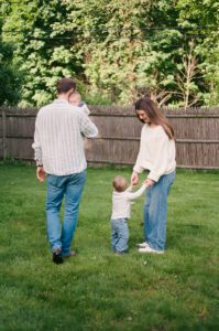 Family of four plays in their backyard in Greater Boston. The mother holds hands with a toddler boy, both wearing white, and the father in a blue striped shirt holds a newborn girl.