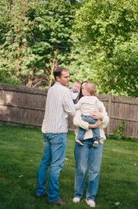 Family of four plays in their backyard in Acton, Massachusetts. The mother holds a toddler boy, both wearing white, and the father in a blue striped shirt holds a newborn girl. Captured on 35mm film.