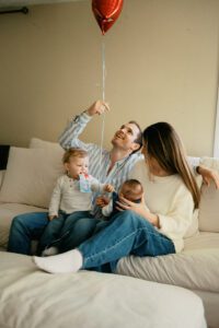 A family plays at home in their living room near Boston. They sit on a white couch, the mother holding a newborn girl while a toddler eats a snack and reaches for his sister's face. The father holds a red balloon.