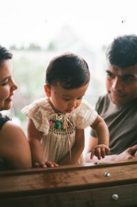 A mother and father look at their one-year-old daughter in a white dress as they sit on a bench in Cambridge, Massachusetts. The father sits next to them smiling while rain pours down behind them. Captured on 35mm film.