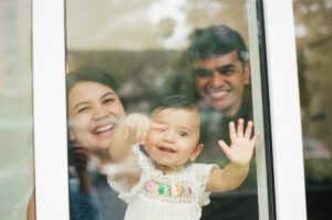 A view through a window of a mother and father smiling behind their one-year-old daughter in a white dress, who is pointing out the window in Cambridge, Massachusetts. Captured on 35mm film.