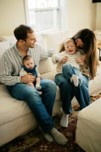 A family laughs and cuddles on a white couch in Littleton, Massachusetts. The mom holds a blonde toddler boy and the dad holds a small newborn girl.