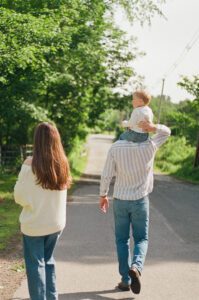 A family walks down the street in Littleton, Massachusetts. A toddler boy sits on his dad's shoulders. They all wear blue and white and jeans. Captured on 35mm film.