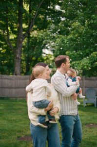 Family of four stands in their backyard looking into the distance in Acton, Massachusetts. The mother holds a toddler boy, both wearing white, and the father in a blue striped shirt holds a newborn girl.