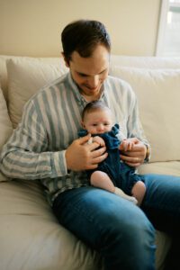 A father in a striped blue shirt holds his newborn daughter in his lap on a white couch in Littleton, Massachusetts.