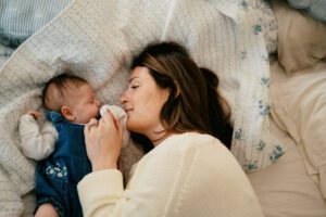 A mother with dark hair and a white sweater snuggles with her sleeping newborn daughter on a bed with blue and white sheets in Littleton, Massachusetts.