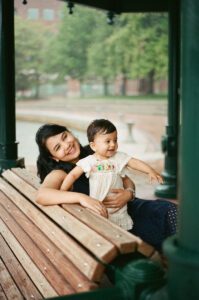 A mother and a toddler girl in a white dress smile on a bench by the Charles river in Cambridge, Massachusetts while rain pours down behind them. Captured on 35mm film.