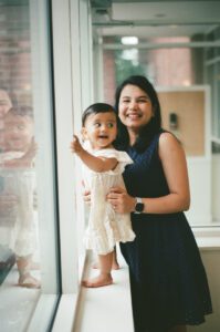 A mother in a dark blue dress stands with her one-year-old daughter in a white dress near the windows of the lobby of their apartment building in Cambridge, Massachusetts. Captured on 35mm film.