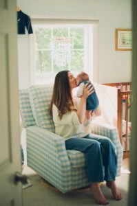 A mother in a white sweater and jeans holds her newborn daughter up and kisses her face. They are a sitting in a blue gingham recliner in a nursery near Acton, Massachusetts.