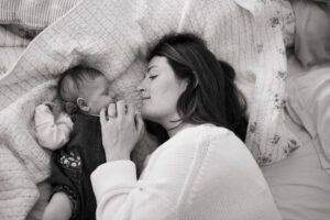 A black and white image of a mother with dark hair and a white sweater snuggles with her sleeping newborn daughter on a bed with blue and white sheets in Littleton, Massachusetts.
