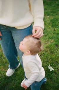 In their backyard in Greater Boston, a woman's arm in a white sweater reaches down touch the head of her toddler boy, both wearing white. Captured on 35mm film.