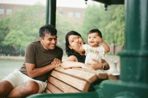 A mother, father, and a toddler girl in a white dress laugh on a bench by the Charles river in Cambridge, Massachusetts while rain pours down behind them. Captured on 35mm film.