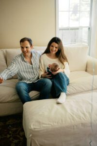 A father in a striped blue sits next to a mother in a white sweater holding their newborn daughter on a white couch in Littleton, Massachusetts.