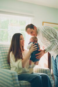 A mother in a white sweater and jeans holds her newborn daughter up as the father leans over them and smiles. They are a sitting in a blue gingham recliner in a nursery near Acton, Massachusetts.