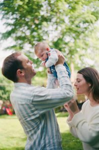 A dad in a blue and white striped shirt lifts his newborn daughter into the air while her mother touches her feet in their backyard in Greater Boston.Captured on 35mm film