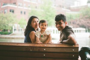 A mother, father, and a toddler girl in a white dress smile on a bench by the Charles river in Cambridge, Massachusetts while rain pours down behind them. Captured on 35mm film.