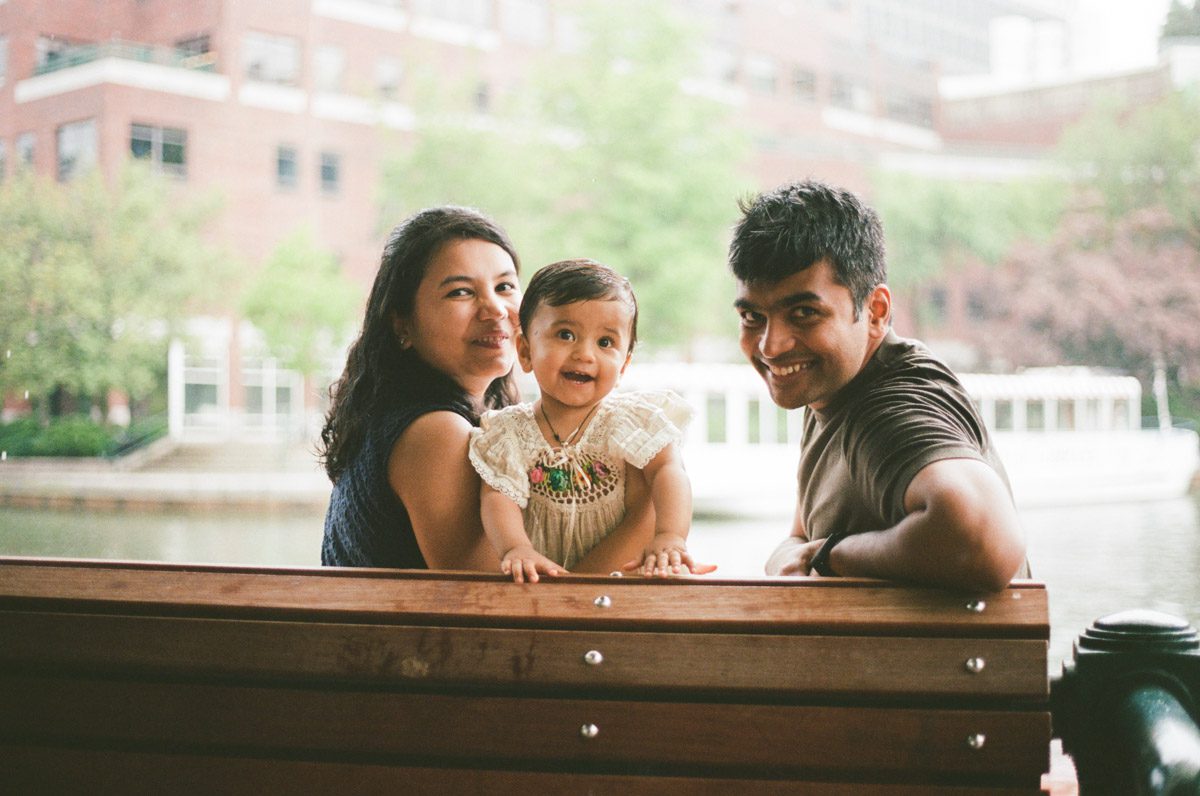 A mother, father, and a toddler girl in a white dress smile on a bench by the Charles river in Cambridge, Massachusetts while rain pours down behind them. Captured on 35mm film.