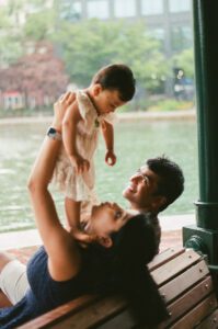 A mother, lifts her one-year-old daughter in a white dress as they sit on a bench by the Charles river in Cambridge, Massachusetts. The father sits next to them smiling while rain pours down behind them. Captured on 35mm film.