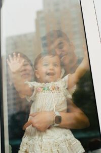 A view through a window of a mother in a dark blue dress holding her one-year-old daughter in a white dress while the father stands behind them and smiles. They are in the lobby of their apartment building in Cambridge, Massachusetts. Captured on 35mm film.