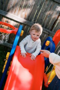 A blonde toddler boy in a white shirt playing on a red slide in his backyard in Acton, Massachusetts. His mother's hand reaches toward him in a white sleeve.
